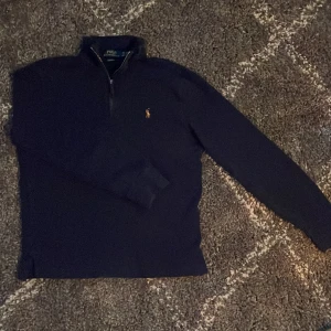 Mörkblå stickad tröja Polo Ralph Lauren - Mörkblå stickad tröja från Polo Ralph Lauren med half zip och unik broderad logga på bröstet. Tröjan har lång ärm, ribbade muddar och hög krage. Perfekt för dig som gillar stilren och tidlös design. Köpt för 1800 på hede outlet (kvitto finns) xs men passar s