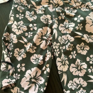Bruna blommiga shorts med bälte - Säljer ett par gröna shorts från Roxy med stora ljusrosa blommor och praktiskt bälte i midjan. Shortsen har fickor både fram och bak och stängs med kardborre. Perfekta för sommaren och strandhäng!