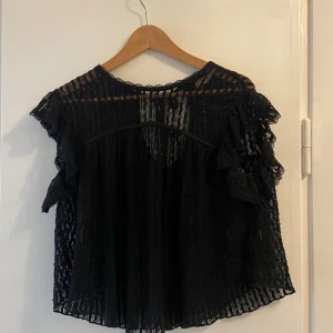 Svart spetsblus med volangdetaljer - Säljer en svart blus i transparent spets med volangdetaljer på ärmarna och knytning i nacken. Blusen har ett luftigt fall och är ärmlös med fina volangiga spetskanter. 