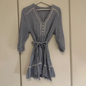Randig blåvit kortklänning från Zara - Superfin blåvit randig klänning från Zara med trekvartsärm, v-ringning och knappar framtill. Klänningen har knytband i midjan och volang nertill för en extra söt look. Perfekt för varma dagar och enkel att styla med sneakers eller sandaler.