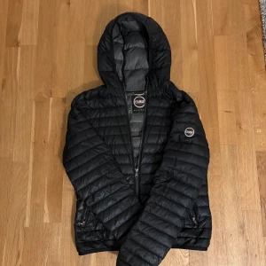 Svart dunjacka med huva från Colmar - Snygg svart dunjacka från Colmar med huva och dragkedja framtill. Jackan har quiltad design, två fickor med dragkedja och Colmar-logga på ärmen. Perfekt för kyliga dagar och enkel att matcha med allt. Trasig dragkedja (går att stänga som vanligt) pris kan diskuteras 