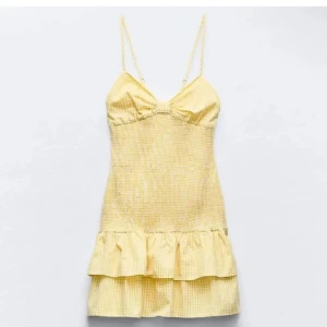 Gul kortklänning med volang - Supersöt gul kortklänning med tunna axelband och volang nertill. Klänningen har smockad överdel och subtilt rutigt mönster. Perfekt för varma dagar och ger en fräsch sommarkänsla. Sitter figurnära upptill och har en lätt utsvängd kjol.