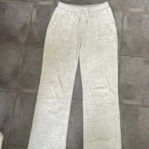 Grå mjukisbyxor med snörning - Snygga ljusgrå mjukisbyxor från Juicy Couture med snörning i midjan och praktiska fickor både fram och bak. Byxorna har raka ben och är gjorda i mjuk bomullsmix, perfekta för chill dagar. JC-logga broderad på ena fickan framtill. Byxorna har 2 små hål på vänster ben. Byxorna har också lite skada runt hålen vid snörena.  !Pris kan diskuteras!