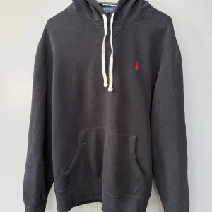 Svart hoodie från Polo Ralph Lauren - Svart hoodie från Polo Ralph Lauren med vit snörning i huvan och röd broderad logga på bröstet. Klassisk känguruficka framtill och mjukt bomullsmaterial. Perfekt för en avslappnad och stilren look.