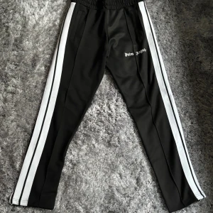 Palm Angels svarta byxor  - Svarta trackpants från Palm Angels med vita sidoränder och broderad logga på låret. Byxorna har elastisk midja, raka ben och dragkedjor vid bensluten. Passar även medium är 182vm lång passar mig utmärkt hänger lite me satt passar även längre.