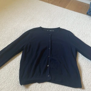 Svart kofta från MNG med knappar - Svart klassisk kofta från MNG i storlek M. Den har rund hals, långa ärmar och stängs med svarta knappar framtill. Materialet är mjukt och stickat, perfekt för lager på lager. Ribbstickade muddar vid ärmslut och nederkant ger en clean look.