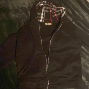 Svart Burberry hoodie med rutigt foder - Svart hoodie från Burberry i storlek M med klassiskt rutigt foder i huvan i svart, vitt och rött. Tröjan har dragkedja framtill, två fickor och långa ärmar. Perfekt för dig som vill ha en stilren och lyxig streetwear-look.