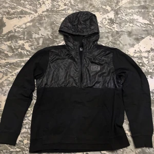 Svart EA7 hoodie med halv dragkedja - Svart hoodie från EA7 Emporio Armani med glansig logomönstrad panel upptill och på huvan. Halv dragkedja framtill, känguruficka och långärmad modell. Materialmix av bomull och syntet, perfekt för en sportig och stilren look.