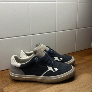Mörkblå sneakers från Pepe Jeans - Snygga mörkblå sneakers från Pepe Jeans med vita detaljer och logga i guld på sidan. Skorna har rund tå, platt sula och snörning. Ovandelen är i mocka och textil, med vit och grå häl samt vit sula. Perfekta för en avslappnad streetstil.