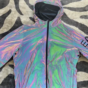 Reflekterande vindjacka från EA7 Armani - Säljer en riktigt fet reflekterande vindjacka från EA7 Emporio Armani. Jackan har huva, hel dragkedja och stort EA7-tryck på ärmen. Materialet är lätt och glansigt, med en galen regnbågseffekt i ljuset. Perfekt för dig som vill sticka ut och gillar streetstyle.