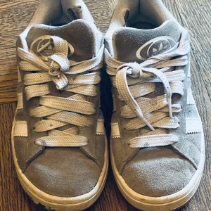 Grå Adidas Campus sneakers i mocka - Snygga Adidas Campus sneakers i grå mocka med vita läderdetaljer och klassiska tre ränder på sidan. Skorna har rund tå, platt sula och vita snören. Adidas logga syns både på plös och häl. Perfekta för en avslappnad streetstyle.