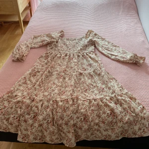 Blommig långklänning med volang - Romantisk långklänning med blommigt mönster i rosa och beige toner. Klänningen har långärmade puffärmar, smockad överdel och volangdetaljer nedtill. Perfekt för dig som gillar en feminin och bohemisk stil. Materialet känns lätt och luftigt.