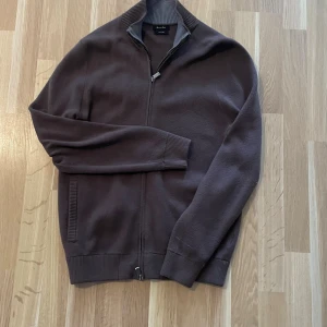 Brun kofta med dragkedja från Massimo Dutti - Mörkbrun kofta från Massimo dutti mjuk stickad bomull. Tröjan har hel dragkedja framtill, ribbade muddar och ståkrage. Perfekt för lager-på-lager och enkel att matcha med jeans eller chinos.