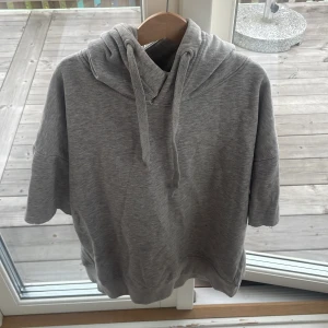 Grå hoodie med kort ärm från lager 157 - Snygg grå hoodie med korta ärmar och huva, från laget 157. Tröjan har gott sönder vid halsen. Gjord i mjuk bomullsmix. Perfekt för en chill och avslappnad stil. Passar dig som gillar streetwear och enkel design.