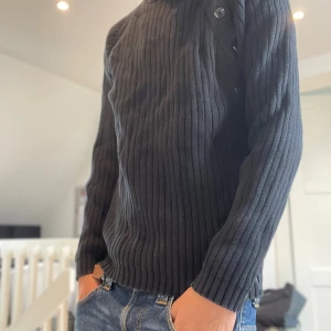 Svart ribbad stickad tröja Jack & Jones - Svart stickad tröja från Jack & Jones med ribbat mönster och coola knappdetaljer på axeln. Långärmad modell med förstärkta armbågar och tryckdetalj nertill. Perfekt för dig som gillar en stilren men ändå unik look.