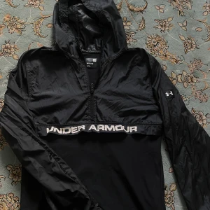 Svart vindjacka från Under Armour - Svart vindjacka från Under Armour med huva och halv dragkedja. Jackan har logga på ärmen och stor vit text över bröstet. Lätt och smidig modell i syntetmaterial, perfekt för aktiva dagar. Snygg sportig look med lång ärm och relaxed fit.
