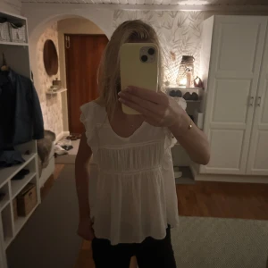 Vit blus med volang från Hunkydory - Supersöt vit blus från Hunkydory med volangdetaljer på axlarna och v-ringning. Blusen har korta ärmar och är tillverkad i ett lätt och luftigt material, perfekt för varmare dagar. Fina sömmar och knapp i nacken ger en extra touch.