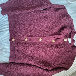 Vinröd kofta från H&M med guldknappar - Vinröd kofta från H&M i storlek S, med snyggt strukturstickat material och sex dekorativa guldknappar framtill. Koftan har långa ärmar och ribbade muddar vid hals och ärmslut. Perfekt för dig som gillar detaljer och vill ha en mysig look. Ordinarie pris:500kr.