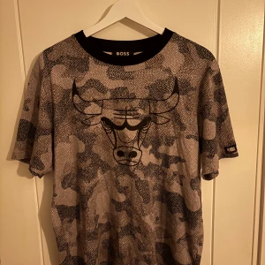 Chicago Bulls x BOSS camo t-shirt - Chicago Bulls x BOSS t-shirt med grått och svart camouflage-mönster, stor bull-logga framtill och svart ribbad krage. Kortärmad och med NBA-märke på ärmen. Perfekt för dig som gillar streetwear och basket. Skick 9/10 hör av er vid frågor 