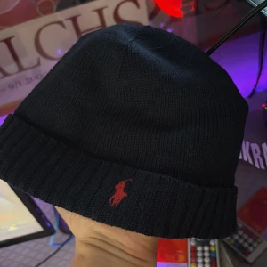 Mörkblå stickad mössa Polo Ralph Lauren - Snygg mörkblå stickad mössa från Polo Ralph Lauren med klassisk röd broderad logga framtill. Mössan har uppvikt kant med ribbad struktur och är tillverkad i mjukt material som håller dig varm. Perfekt för kalla dagar och enkel att matcha med din stil. Priset går självklart att diskutera 