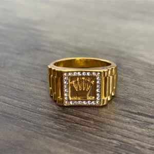 Ring med krona och stenar - Snygg och chunky guldfärgad ring med fyrkantig front, likt en klackring, dekorerad med en krona i mitten och gnistrande vita gnistrande stenar runt om. Ringen har en bred, länk-inspirerad design som ger en lyxig vibe och sticker ut på handen. För män, ungefär storlek 6-7.  