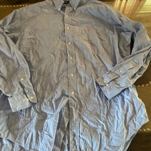 Blå skjorta från Ralph Lauren - Snygg blå skjorta från Ralph Lauren med klassisk button-down krage och broderad logga på bröstet. Tillverkad i 100% bomull, modellen Yarmouth. Skjortan har långa ärmar och vita knappar framtill.