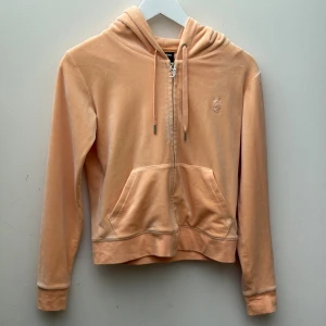 Persikofärgad Juicy Couture hoodie - Supermjuk och lyxig persikofärgad hoodie från Juicy Couture i velour. Tröjan har huva, dragkedja med snygg J-detalj, känguruficka och långa ärmar. Perfekt för en chill och trendig look.  Storlek S i tröjan och storlek XS i byxorna.