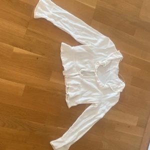 Vit långärmad blus med volang - Supersöt vit blus från H&M med långa ärmar och volangdetaljer vid halsringning och nederkant. Toppen har knyt framtill och är croppad, vilket ger en trendig look. Materialet känns mjukt och lätt, perfekt för vår och sommar.