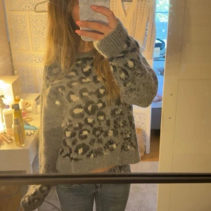 Grå leopardmönstrad stickad tröja - Supermysig grå stickad tröja med leopardmönster i svart och vitt. Tröjan har en loose passform, rund halsringning och långa ärmar med breda muddar. Perfekt för kyliga dagar när du vill vara både snygg och bekväm.