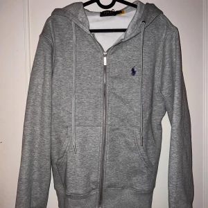 Grå hoodie från Polo Ralph Lauren - Snygg grå hoodie från Polo Ralph Lauren med dragkedja framtill och klassisk blå logga på bröstet. Tröjan har huva med snörning och två fickor framtill. Perfekt för en chill och avslappnad stil.