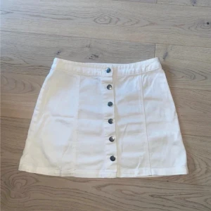 Vit kortkjol med knappar framtill - Säljer en vit kortkjol med snygga silverfärgade knappar längs hela framsidan. Kjolen har en A-linjeformad siluett och är gjord i ett stadigt bomullsmaterial. Perfekt för dig som gillar en clean och fräsch look med lite edge.