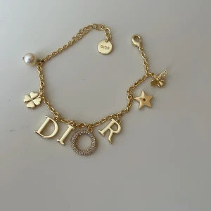 Guldigt Dior armband med berlocker - Supersnyggt guldigt armband från Dior med berlocker som bokstäver, stjärna, fyrklöver, pärla och en liten bi. O:et i Dior är täckt av glittrande stenar för extra bling. Perfekt statement piece med lyxig vibe och coola detaljer. 