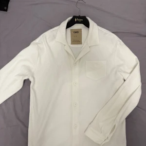 Vit frottéskjorta från OAS, stl S - Vit långärmad skjorta från OAS i mjuk frotté-bomull. Klassisk krage, bröstficka och knäppning framtill. Perfekt för chill dagar eller strandhäng. Enkel och clean design med relaxed vibe.