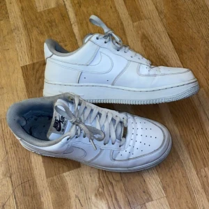Vita Nike Air Force 1 sneakers - Klassiska vita Nike Air Force 1 sneakers med lågt skaft och snörning. Skorna har en rund tå, perforerade detaljer på tån och är tillverkade i skinn med grå insida. Perfekta för en clean och tidlös streetstil.