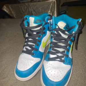 Nike Dunk High blå och vit med neon - Nike Dunk High sneakers i blått och vitt med neonfärgad Swoosh på sidorna. Skorna har svart snörning, perforerad tå och hög siluett. Yttermaterialet är skinn och syntet, med svart platt sula. Perfekt för dig som gillar färgstarka och sportiga sneakers.