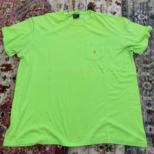 Limegrön t-shirt från Polo Ralph Lauren - Fräsch limegrön t-shirt från Polo Ralph Lauren med klassisk rund hals och korta ärmar. På bröstet finns en ficka med orange broderad Polo-logga. T-shirten är gjord i mjuk bomull och har en loose passform som är chill och bekväm.