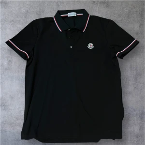 Svart Moncler pikétröja - Storlek M-S. Svart pikétröja från Moncler med korta ärmar och klassisk krage. Tröjan har snygga rödvita ränder på krage och ärmslut samt Moncler-logga på bröstet. Tillverkad i mjuk bomull som känns skön mot huden. Perfekt för dig som gillar stilren design med en sportig touch.