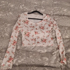 Vit blommig croppad långärmad topp - Superfin croppad topp i vit med blommigt mönster i rosa och rött. Långärmad modell med ribbad struktur och rundad halsringning. Tillverkad i 100% polyester, perfekt för dig som gillar romantiska och söta vibes.🎀