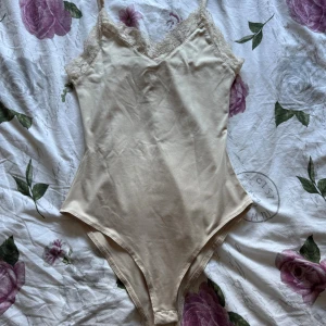 Beige spetsbody från H&M XS - Säljer en beige body från H&M i storlek XS. Den har tunna axelband, v-ringning och fina spetsdetaljer längs urringningen. Materialet är mjukt och stretchigt, perfekt för lager-på-lager eller som statement-piece. Enkel och stilren design. Den ser mycket mer mörkare på bild men är egentligen lite ljusare