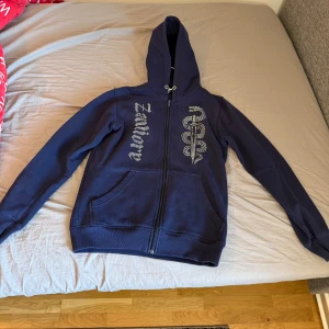 Mörkblå hoodie från Zantiore med tryck - Mörkblå hoodie från Zantiore i storlek S. Tröjan har dragkedja, huva och två fickor framtill. På bröstet finns ett coolt ormmönstrat tryck och texten 'Zantiore' i silver. Perfekt för dig som gillar streetstyle och vill sticka ut.