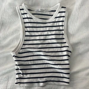Randig croppad linnetopp från Zara - Säljer en fräsch croppad linnetopp från Zara i storlek S. Toppen är vit med svarta horisontella ränder och har ribbad struktur. Modellen är ärmlös med breda axelband och rund halsringning, perfekt för en chill och stilren look. Använd några få gånger för den är för liten.