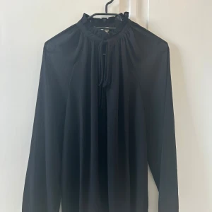 Svart blus - Tunn svart blus. Snören framtill som gör att man kan välja att antingen knyta en rosett eller ha blusen öppen. Tunt luftigt material. Bara använd en gång, nyskick!