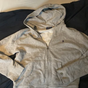 Grå hoodie från Polo Ralph Lauren - Snygg grå hoodie från Polo Ralph Lauren med klassisk blå broderad logga på bröstet. Tröjan har huva, dragkedja framtill och två stora fickor. Perfekt för chill dagar och enkel att matcha med jeans eller joggers.