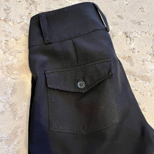 Svarta bootcut-byxor från Gina Tricot - Svarta bootcut-byxor från Gina Tricot med hög midja och snygg passform. Byxorna har ficka med lock och knapp bak samt bälteshällor och dubbla knappar fram. Perfekta för en clean och stilren look.