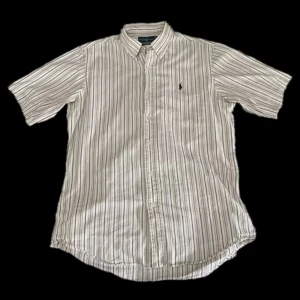 Kortärmad Ralph Lauren skjorta - Kortärmad skjorta från Ralph Lauren i vitt med  ränder. Klassisk krage, knäppning framtill och broderad logga på bröstet. Skjortan har en avslappnad passform, perfekt för en stilren look. (Finns en liten lagning i högra armhålan)