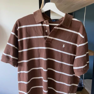 Brun randig polo från Ralph Lauren - Säljer en brun och vit randig pikétröja från Ralph Lauren med klassisk krage och knappar framtill. T-shirten har korta ärmar och är tillverkad i bomull. Perfekt för en chill och stilren look.