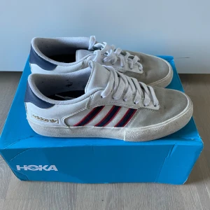 Adidas sneakers med röda och blå ränder - Snygga Adidas sneakers i vitt mocka och textil med klassiska tre ränder i rött och blått på sidorna. Skorna har vit snörning, mörkblå häl och gummisula med mönster. Perfekta för dig som gillar retrostil och streetwear.