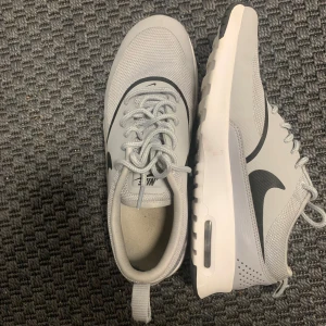 Grå Nike air Max thea sneakers med svart swoosh  - Snygga grå Nike sneakers med svart swoosh på sidan. Ovandelen är i mesh och syntet för en sportig look och bra andningsförmåga. Skorna har snörning, vit sula med synlig luftbubbla och svarta detaljer på yttersulan. Perfekta för en clean och modern stil. Hör av er vid fundering🙌 endast använd 1-2ggr