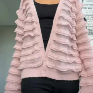 Rosa kofta med volangdetaljer - Superfin ljusrosa kofta med volangdetaljer över hela tröjan. Koftan har knappar framtill och är stickad i ett mjukt material. Perfekt för dig som vill ha en söt och unik look med extra detaljer på ärmar och kropp.