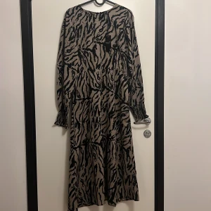 Zebramönstrad långklänning från 157 - Cool långklänning från 157 med zebramönster i svart och beige. Klänningen har långa ärmar med smock vid handlederna och en avslappnad passform. Perfekt för dig som vill sticka ut med ett djurmönster och ändå känna dig bekväm.